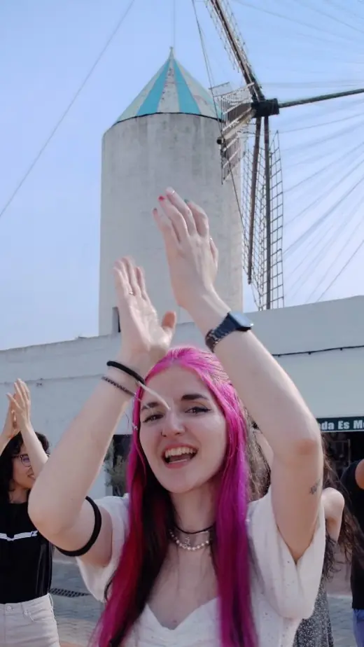 Darlie Ole con i capelli rosa ride a braccia alzate davanti a un mulino bianco